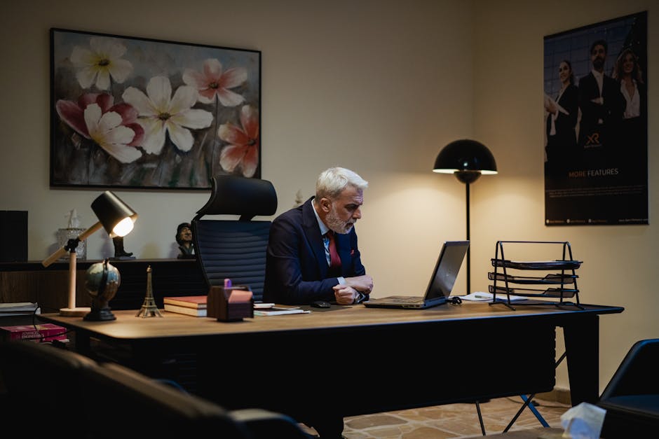A professional man with gray hair and a beard, dressed in a navy suit, is seated at a dark wooden desk in an office, working on a silver laptop. The desk is neatly arranged with a small vintage-style