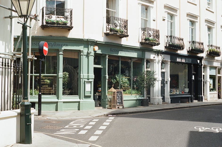 A charming florist shop with a light green facade and large glass windows showcasing fresh flower arrangements. The shop has black wrought-iron balconies above, adorned with small potted plants. Outsi