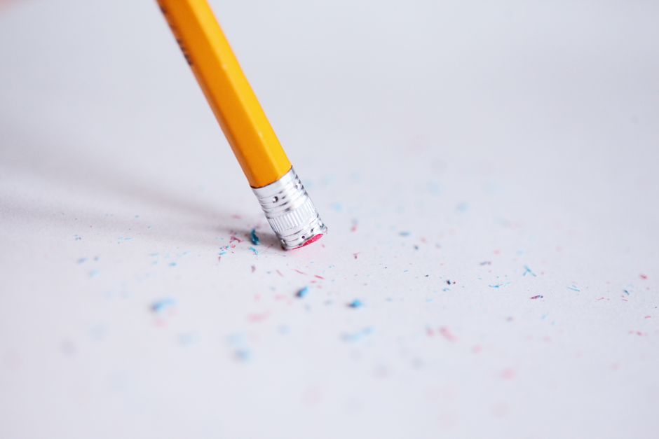 A close-up photograph of a yellow wooden pencil with a silver ferrule and pink eraser, positioned diagonally on a white surface, with colorful shavings and tiny bits of confetti-like debris scattered