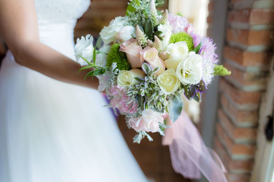 A close-up of a bridal bouquet held by a woman in a white wedding dress. The bouquet features cream and pale pink roses, white lisianthus, and soft purple and green foliage, arranged in a rounded styl