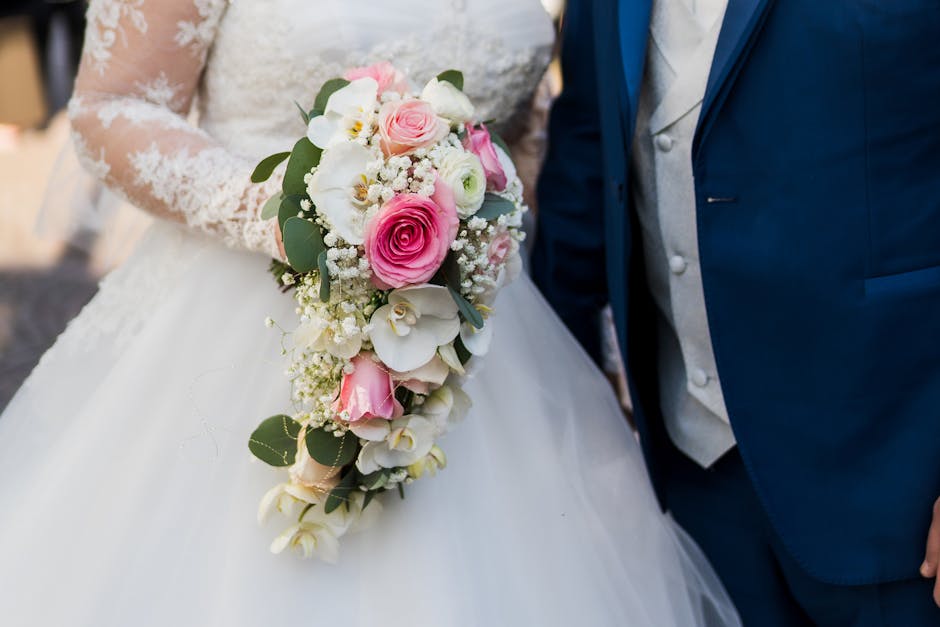 A close-up of a wedding floral bouquet featuring pink roses, white orchids, and small white filler flowers, accented with green eucalyptus leaves, held by a bride dressed in a lace wedding gown. The b