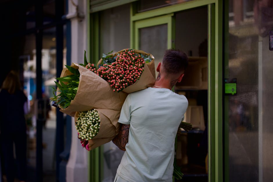 A man with short dark hair and a tattooed arm is carrying a large bouquet of fresh flowers wrapped in brown paper outside a florist shop with a green doorframe. The bouquet includes pink and white flo