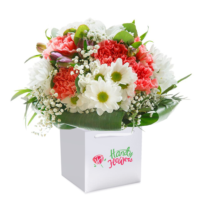 Arranged in a contemporary square white gift bag, this lush bouquet encapsulates the elegance and diversity of West Kensington. Ruffled petals of coral and peach carnations bring texture and an inviting softness, while daisy-like white chrysanthemums with vivid green eyes pop with crisp, joyful contrast. Airy tendrils of baby's breath scatter a subtle lightness among the larger blooms. A few tight, purple-tinted buds are visible, providing layers of colour and intrigue reminiscent of a stroll along the vibrant shops of North End Road. Deep, glossy green foliage shapes the base, its curved leaves wrapping the bouquet in a lush collar that nods to the classic gardens hidden throughout West Kensington. The rounded, full silhouette reflects modern sensibilities while honouring timeless design. The bag's sturdy white ribbon handles add grace and easy portability, symbolising the convenience of local, on-demand delivery to homes from Barons Court to Kensington Olympia. Every detail-from the premium flower selection to the careful placement of each stem-reflects the skill, heart, and neighbourhood pride that sets West Kensington Florist apart.